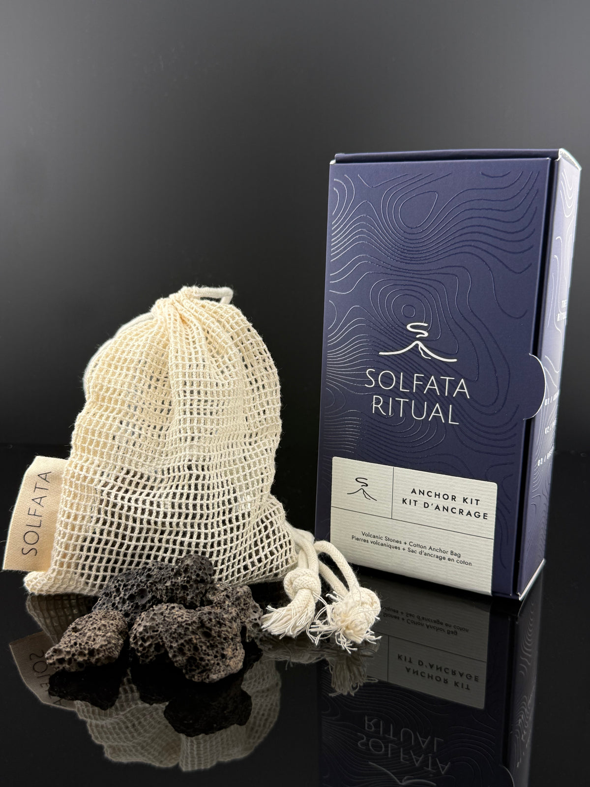 Solfata Ritual anchor kit featuring a navy blue box, a cream mesh drawstring bag, and dark volcanic pumice stones on a reflective black surface.