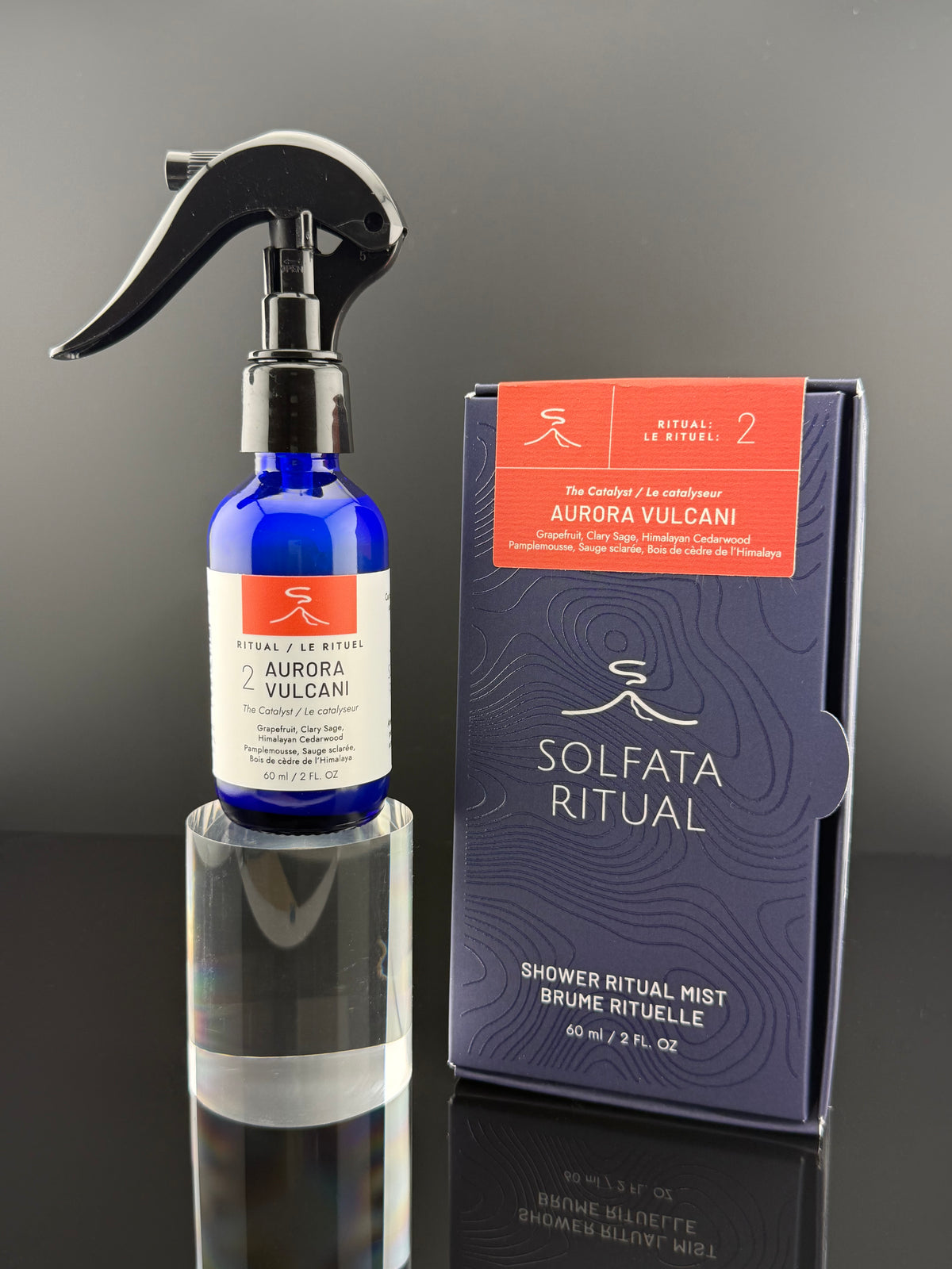 A cobalt blue glass shower ritual mist bottle with a black spray trigger and white label, standing beside its dark navy and orange box on a reflective surface.