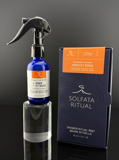 A cobalt blue spray bottle with a black trigger sprayer stands on a reflective clear pedestal next to its dark navy and orange retail box. The label reads 'Solfata Ritual' and 'Ignis et Rosa'.